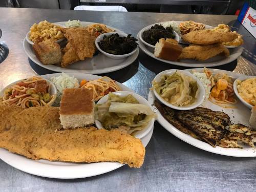 Two plates of Southern-style food featuring fried fish, macaroni, greens, coleslaw, and cornbread on a metal table.