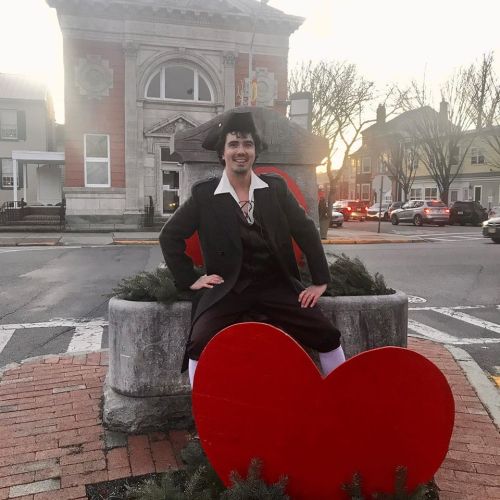 A person in historical attire sits on a large red heart sculpture in a town square, with a building in the background.