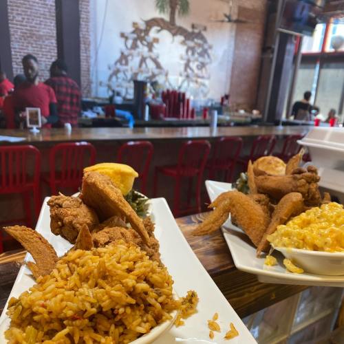 Two plates of fried chicken with rice and macaroni, set against a restaurant backdrop with a bar and patrons.