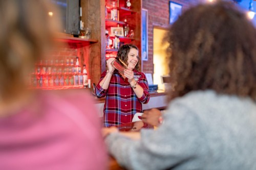 A bartender shakes a cocktail while smiling at two customers seated at the bar. The setting is warm and inviting.