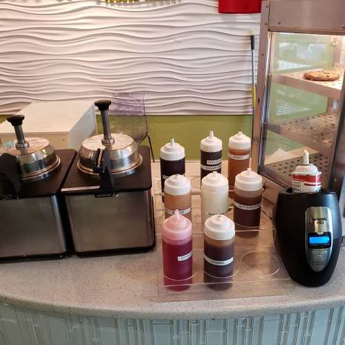 Two waffle makers and a variety of squeeze bottles filled with sauces on a countertop. A display case is in the background.