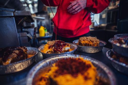 A person in a red hoodie drizzles sauce over trays of colorful, topped dishes in a food stall.
