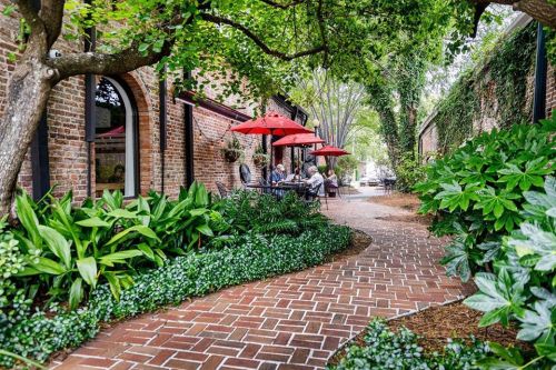 A brick pathway leads through lush greenery to a cozy outdoor dining area with red umbrellas.