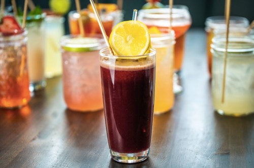 A vibrant drink with a lemon slice, surrounded by various colorful cocktails in jars on a wooden table.