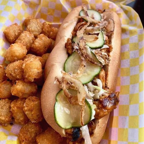 A hot dog topped with sliced cucumbers and crispy onions, served with a side of golden tater tots in a checkered basket.
