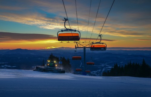 A ski lift with orange chairs against a sunset backdrop, with a snow groomer preparing the slope below.