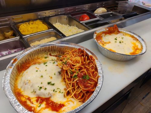 Two aluminum trays filled with spaghetti and baked cheese, garnished with parsley, in a kitchen setting.