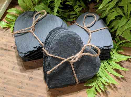 A stack of heart-shaped slate coasters tied with twine, surrounded by green ferns on a wooden surface.