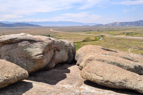 A rocky outcrop overlooks a vast, open landscape with a winding road and distant mountains under a blue sky.