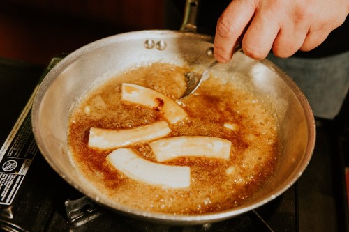 A hand stirs frying pieces of food in a pan with bubbling oil on a stovetop.