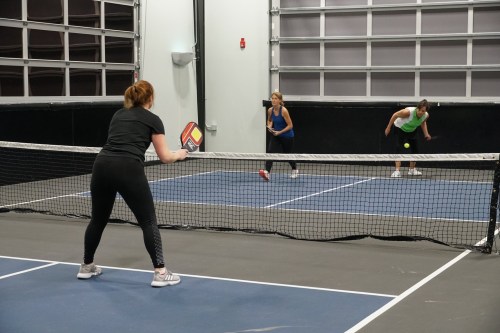 Two women play pickleball while a third woman prepares to serve, all on an indoor court with a net.