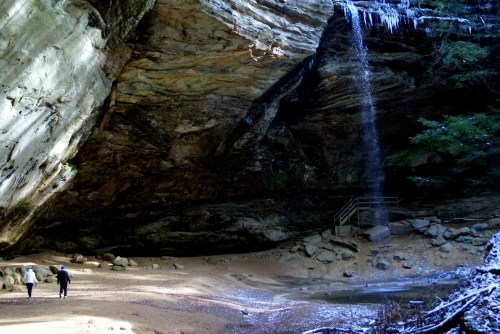 Two people walk along a sandy area beneath a large rock formation with a waterfall and icicles in a forested setting.