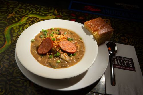 A bowl of gumbo with sausage slices, topped with green onions, served with cornbread on the side.