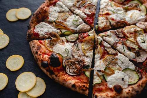 A freshly baked pizza topped with cheese, zucchini, and onions, sliced into wedges, with yellow squash slices nearby.