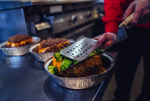 A person uses a spatula to add broccoli and cheese to a baked dish in a kitchen, with other dishes in the background.