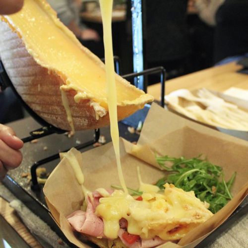 Melted cheese being poured over a sandwich with ham, tomatoes, and arugula on a wooden table.