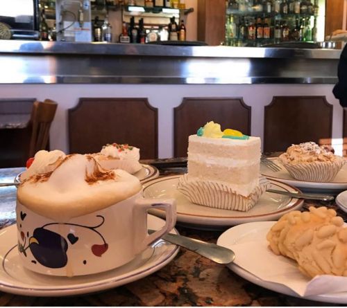 A cozy café table with a frothy cappuccino, a slice of white cake, and assorted pastries in the background.
