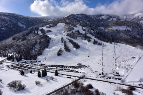 A snowy mountain landscape with ski slopes, trees, and a parking lot filled with cars under a blue sky.
