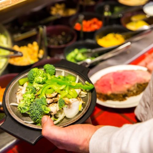 A person holds a plate of stir-fried vegetables, including broccoli and green peppers, in front of a buffet.