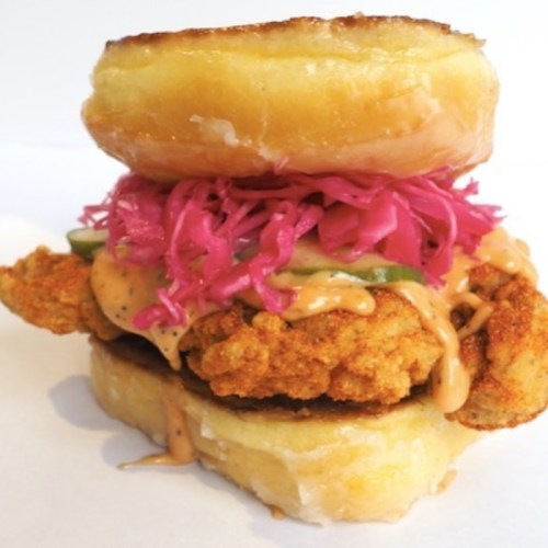 A stacked sandwich featuring a fried chicken fillet, pickled vegetables, and spicy sauce between two glazed donuts.