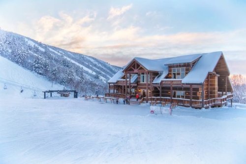 A cozy wooden lodge surrounded by snow-covered mountains and a ski lift, under a colorful sky at sunset.