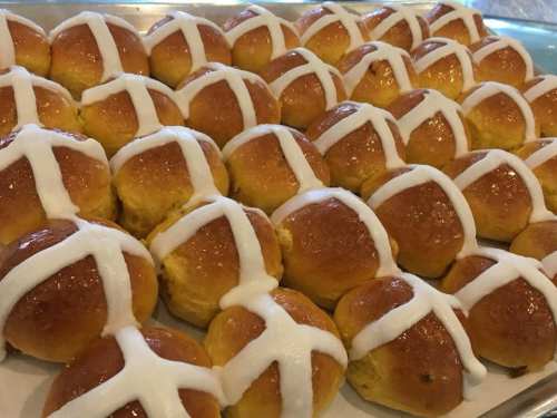 A close-up of freshly baked hot cross buns, glazed and decorated with white icing crosses on top.