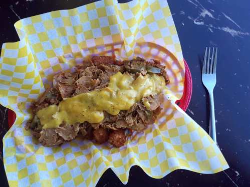 A plate of crispy tater tots topped with shredded meat and drizzled with yellow mustard, served in a checkered paper.