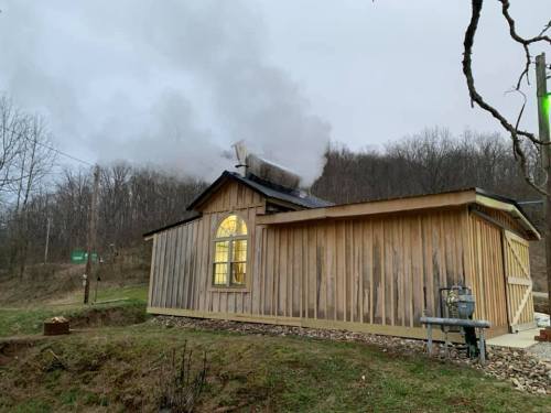 A wooden cabin with smoke rising from the chimney, set against a backdrop of trees and a cloudy sky.