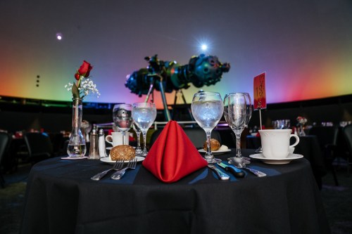 A beautifully set dining table with elegant glassware, a red napkin, and a rose, against a colorful backdrop.