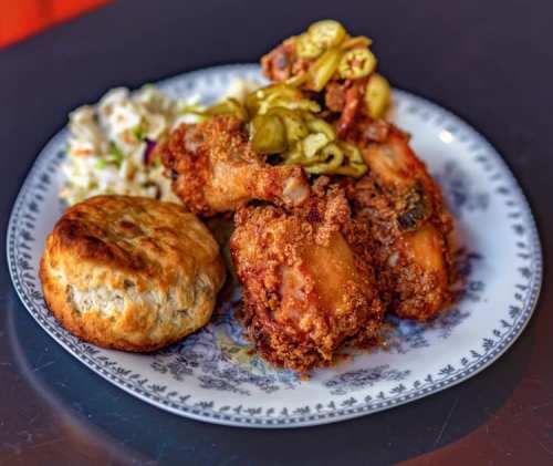 A plate of fried chicken with pickles, coleslaw, and a biscuit, served on a decorative blue and white dish.
