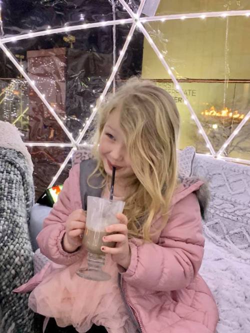 A young girl with long blonde hair enjoys a drink while sitting in a cozy, decorated space with lights.