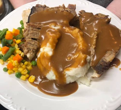 A plate of sliced roast beef with gravy, mashed potatoes, and a side of mixed vegetables including carrots, peas, and corn.