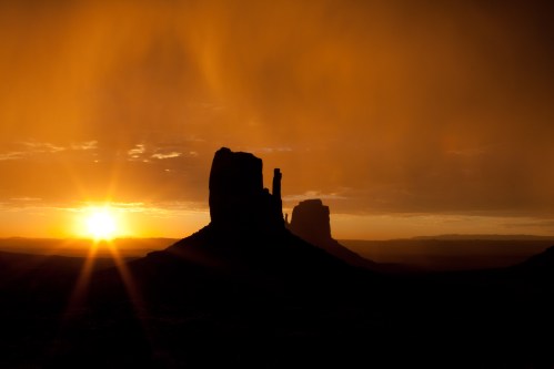 Silhouetted rock formations against a vibrant orange sunset, with dramatic clouds in the sky.