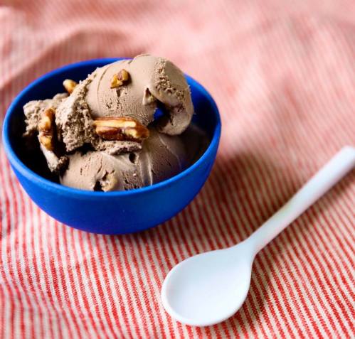 A blue bowl filled with chocolate ice cream topped with nuts, placed on a red and white striped cloth with a white spoon.