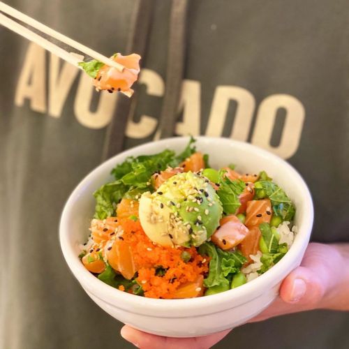 A person holding a bowl of sushi rice topped with salmon, avocado, and vegetables, with chopsticks in hand.