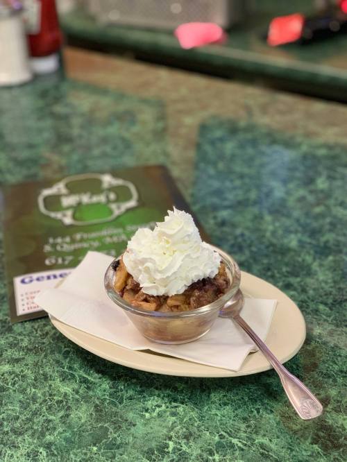 A glass dish of dessert topped with whipped cream, sitting on a plate with a spoon, on a green countertop.