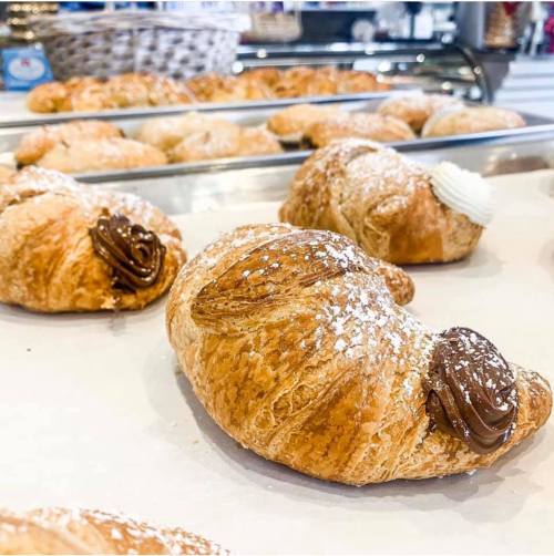 A close-up of a flaky croissant filled with chocolate spread, dusted with powdered sugar, on a tray with more pastries.