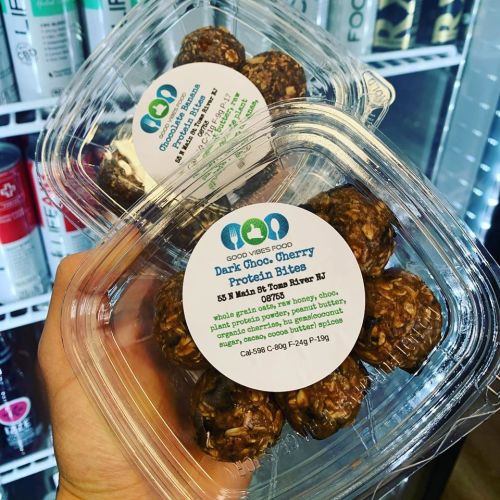 A hand holding two containers of dark chocolate cherry protein bites with labels detailing ingredients and nutritional info.