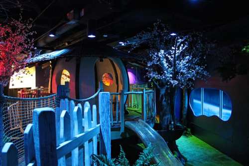 A colorful indoor play area featuring a treehouse, slide, and vibrant lighting with decorative trees and a fence.