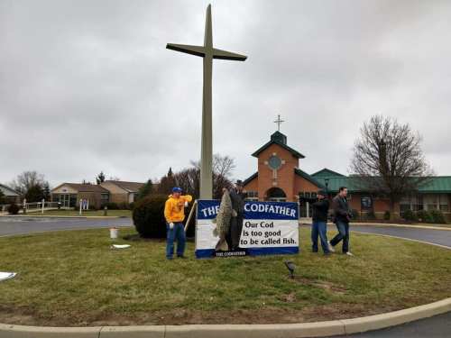 Three people stand by a large cross outside a church, holding a humorous sign about cod and fish. Overcast sky.