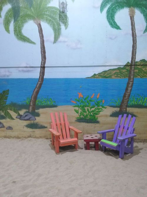 Two colorful adirondack chairs, pink and purple, sit on sandy ground with a tropical mural of palm trees and ocean in the background.