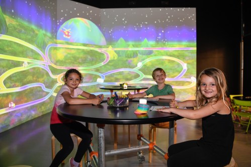 Three children sit at a table in a colorful, interactive room with vibrant projections on the walls.