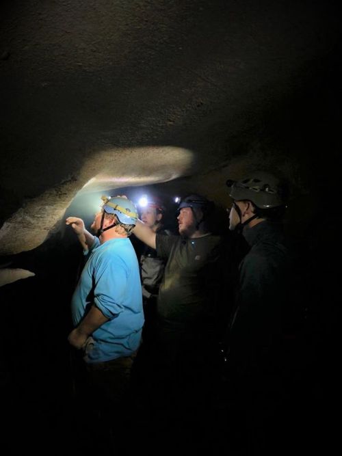 A group of four people wearing helmets explores a dark cave, illuminated by headlamps.