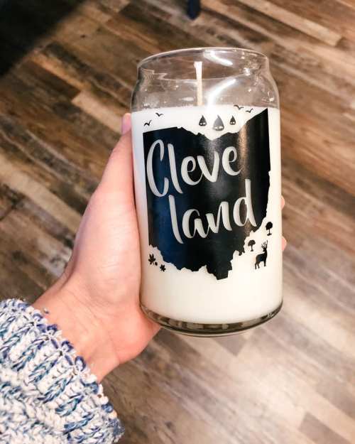 A hand holding a candle in a glass jar with a design of Ohio and the word "Cleveland" in bold letters.
