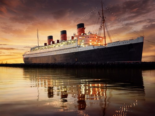 The Queen Mary ship at sunset, illuminated with lights, reflecting in calm waters.