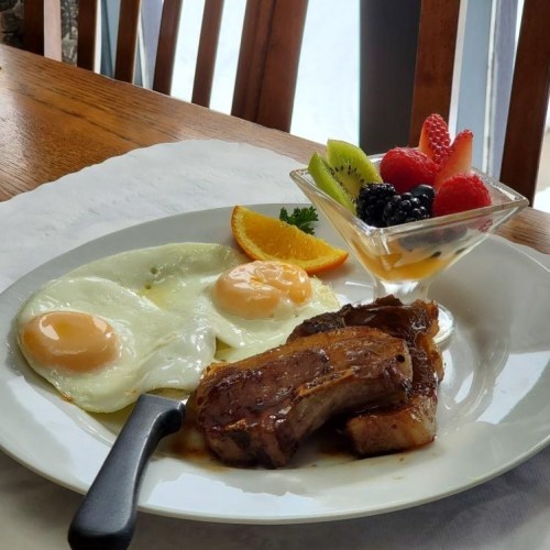 A plate with two fried eggs, grilled meat, and a fruit cup with berries and kiwi, set on a wooden table.