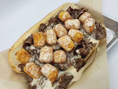 A loaded sandwich with sliced beef, tater tots, and creamy sauce on a toasted hoagie roll, served on parchment paper.