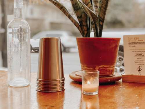 A wooden table with a glass bottle, metal cups, a candle, a plant, and a menu in a cozy café setting.