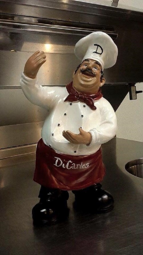 A cheerful chef figurine wearing a white hat and apron, gesturing as if welcoming guests in a kitchen setting.
