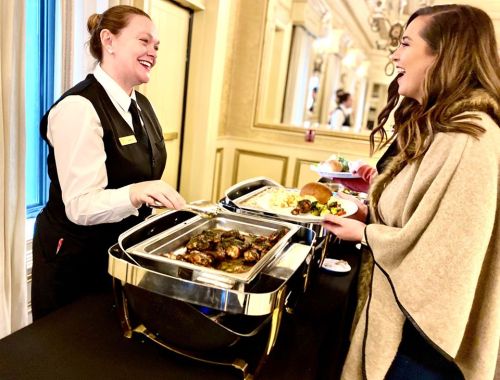 A smiling server in a black vest serves food to a woman in a beige shawl at a buffet in an elegant setting.